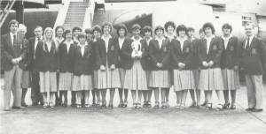 A group of about 20 women stand in front of an airplane wearing dowdy skirt suits