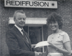 June Jaycocks smiling outside Rediffusion's building, shaking hands with and accepting a cheque from the company's manager