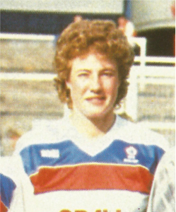 Head and shoulders shot of a smiling young woman with short curly brown hair in a white t shirt with red and blue stripes