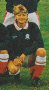 A crouching smiling blond haired woman in a football kit with red socks, white shorts, a blue sweatshirt and a blue headband