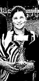 A smiling young woman in a football shirt being presented with a trophy.